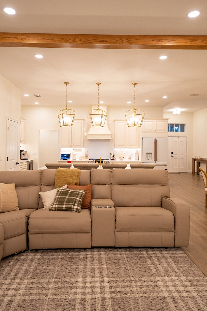 Open-concept living room featuring a beige reclining sectional sofa, plaid area rug, and a white kitchen with gold lantern pendant lights.