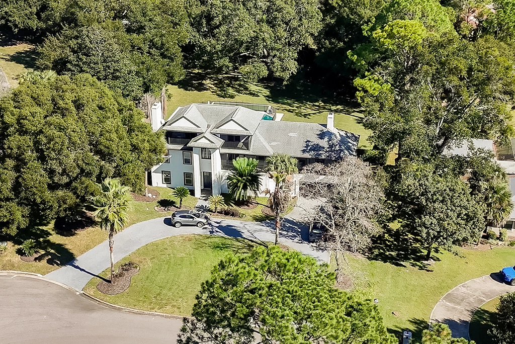 Aerial view of a luxury white estate home featuring a grand entrance, circular paved driveway, and lush landscaping with mature oak trees.